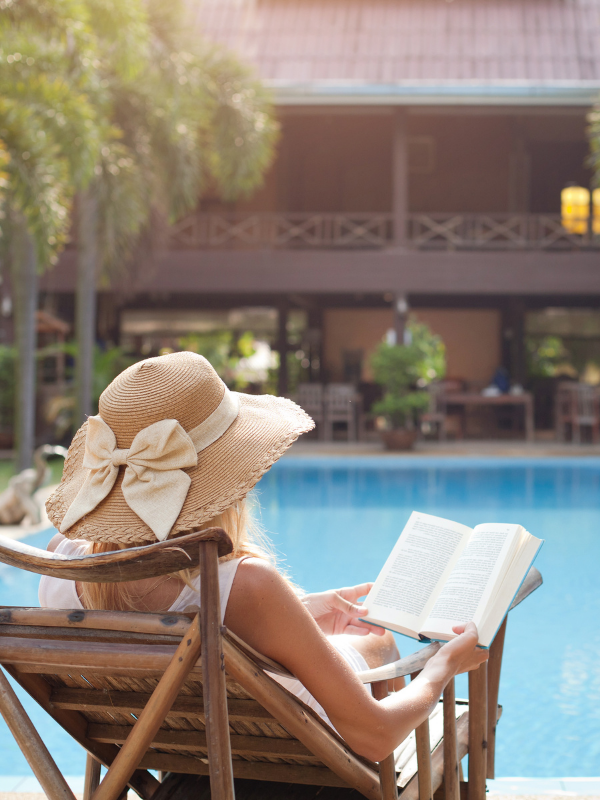 A woman is in a poolside lounge chair in her sunhat reading a book. 